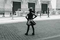 A bronze statue of a young girl with her hands on her hips stands confidently on a cobblestone street. She faces a building with ornate architecture and closed shutters, separated by a black iron fence. The image conveys a sense of empowerment through the girl's determined stance.