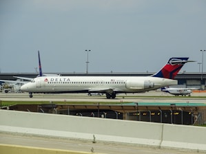 A white Delta Airlines aircraft is stationed on the ground at an airport. Additional infrastructure and smaller aircraft are visible in the background. The sky appears overcast.