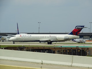A white Delta Airlines aircraft is stationed on the ground at an airport. Additional infrastructure and smaller aircraft are visible in the background. The sky appears overcast.
