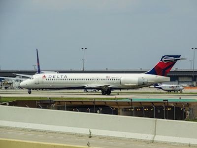 A white Delta Airlines aircraft is stationed on the ground at an airport. Additional infrastructure and smaller aircraft are visible in the background. The sky appears overcast.