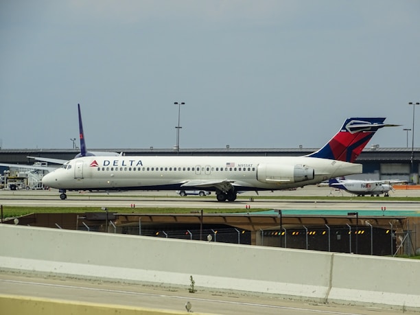 A white Delta Airlines aircraft is stationed on the ground at an airport. Additional infrastructure and smaller aircraft are visible in the background. The sky appears overcast.