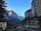 Charming alpine village surrounded by towering cliffs and a cascading waterfall. Traditional wooden buildings with a backdrop of snow-capped mountains create a picturesque scene. Swiss flags are visible, adding a cultural touch to the serene landscape.