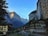 Charming alpine village surrounded by towering cliffs and a cascading waterfall. Traditional wooden buildings with a backdrop of snow-capped mountains create a picturesque scene. Swiss flags are visible, adding a cultural touch to the serene landscape.
