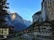 Charming alpine village surrounded by towering cliffs and a cascading waterfall. Traditional wooden buildings with a backdrop of snow-capped mountains create a picturesque scene. Swiss flags are visible, adding a cultural touch to the serene landscape.