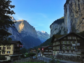 Charming alpine village surrounded by towering cliffs and a cascading waterfall. Traditional wooden buildings with a backdrop of snow-capped mountains create a picturesque scene. Swiss flags are visible, adding a cultural touch to the serene landscape.