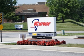 A sign stands prominently near the road, displaying 'US Lacrosse' in large red letters above a logo, and below it reads 'MedStar Health' along with an address '2 Loveton Circle'. The sign is surrounded by well-maintained landscaping, featuring red flowers and neatly trimmed grass. In the background, there is a building with a brick facade, trees, and additional signage, indicating a peaceful, suburban setting.