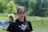 A woman wearing protective eyewear stands outdoors on a sports field. She is dressed in a black shirt with a 'Brave' logo and is holding a lacrosse stick. The background features greenery, indicating a sunny day.