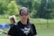 A woman wearing protective eyewear stands outdoors on a sports field. She is dressed in a black shirt with a 'Brave' logo and is holding a lacrosse stick. The background features greenery, indicating a sunny day.