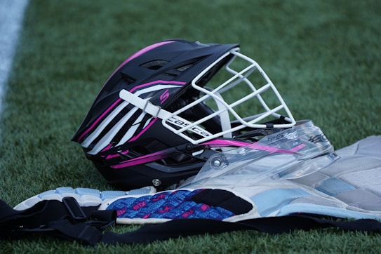 A lacrosse helmet with a black exterior and vibrant pink and white stripes is lying on a patch of green grass. The helmet features a white metal faceguard. Next to it, lacrosse protective gear, including a chest pad with blue and white elements, is partially visible.