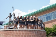 A group of young people wearing matching sports shirts and shorts stand on a circular balcony in front of a brick building. They are smiling and appear to be posing for a photo. Two large statues holding lacrosse sticks are visible behind them. The sky is clear with some clouds, and there is greenery visible at the bottom of the image.