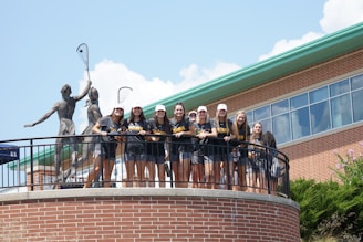 A group of young people wearing matching sports shirts and shorts stand on a circular balcony in front of a brick building. They are smiling and appear to be posing for a photo. Two large statues holding lacrosse sticks are visible behind them. The sky is clear with some clouds, and there is greenery visible at the bottom of the image.
