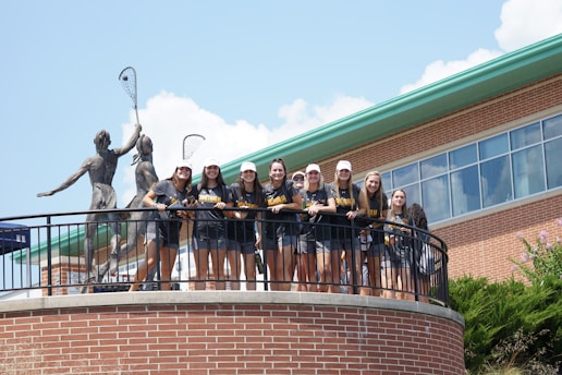 A group of young people wearing matching sports shirts and shorts stand on a circular balcony in front of a brick building. They are smiling and appear to be posing for a photo. Two large statues holding lacrosse sticks are visible behind them. The sky is clear with some clouds, and there is greenery visible at the bottom of the image.
