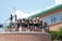 A group of young people wearing matching sports shirts and shorts stand on a circular balcony in front of a brick building. They are smiling and appear to be posing for a photo. Two large statues holding lacrosse sticks are visible behind them. The sky is clear with some clouds, and there is greenery visible at the bottom of the image.