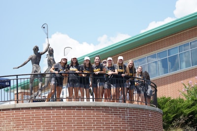A group of young people wearing matching sports shirts and shorts stand on a circular balcony in front of a brick building. They are smiling and appear to be posing for a photo. Two large statues holding lacrosse sticks are visible behind them. The sky is clear with some clouds, and there is greenery visible at the bottom of the image.
