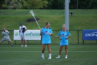 Two women in matching light blue athletic uniforms stand on a sports field, with one holding a lacrosse stick. Other individuals, including a photographer, can be seen in the background. The setting appears to be outdoors with greenery surrounding the area and advertisements on the fence.