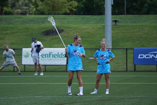 Two women in matching light blue athletic uniforms stand on a sports field, with one holding a lacrosse stick. Other individuals, including a photographer, can be seen in the background. The setting appears to be outdoors with greenery surrounding the area and advertisements on the fence.