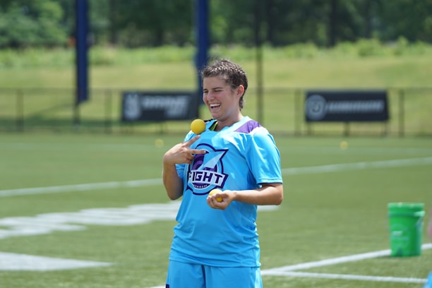 A person in a blue sports jersey stands on a grass field, smiling and holding two yellow balls. A green bucket is visible on the right side, and the background includes a fenced sports ground with blurred trees.