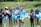 A group of female athletes stand on a sports field, with some clapping and one in the center raising her hand energetically. The players wear sports uniforms, and a few are in casual attire, indicating a mix of team members and perhaps coaching staff. The setting is outdoors, with a grassy background and a sunny atmosphere.
