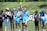 A group of female athletes stand on a sports field, with some clapping and one in the center raising her hand energetically. The players wear sports uniforms, and a few are in casual attire, indicating a mix of team members and perhaps coaching staff. The setting is outdoors, with a grassy background and a sunny atmosphere.