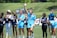 A group of female athletes stand on a sports field, with some clapping and one in the center raising her hand energetically. The players wear sports uniforms, and a few are in casual attire, indicating a mix of team members and perhaps coaching staff. The setting is outdoors, with a grassy background and a sunny atmosphere.