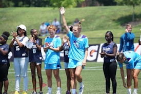 A group of female athletes stand on a sports field, with some clapping and one in the center raising her hand energetically. The players wear sports uniforms, and a few are in casual attire, indicating a mix of team members and perhaps coaching staff. The setting is outdoors, with a grassy background and a sunny atmosphere.