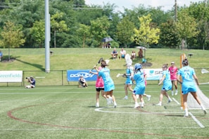 A group of athletes in blue uniforms are playing a game of lacrosse on a grassy field. Some are wearing helmets and holding lacrosse sticks, while others wear pink referee shirts. A small audience sits on a hill in the background, surrounded by trees. Advertising banners line the fence next to the field.