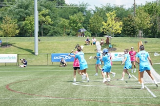 A group of athletes in blue uniforms are playing a game of lacrosse on a grassy field. Some are wearing helmets and holding lacrosse sticks, while others wear pink referee shirts. A small audience sits on a hill in the background, surrounded by trees. Advertising banners line the fence next to the field.