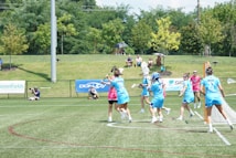 A group of athletes in blue uniforms are playing a game of lacrosse on a grassy field. Some are wearing helmets and holding lacrosse sticks, while others wear pink referee shirts. A small audience sits on a hill in the background, surrounded by trees. Advertising banners line the fence next to the field.