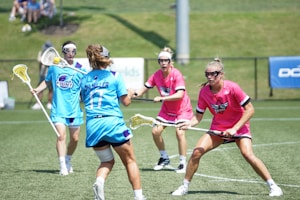 Four female lacrosse players are actively engaged in a game on a grassy field. Two players are wearing blue uniforms with one in possession of the ball, ready to pass or shoot, while two others in pink uniforms are defending against them. All players are wearing protective eyewear, and the background shows a fence and spectators seated on a hill.