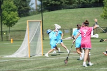 A group of female lacrosse players on a field, with one wearing a pink jersey and others in blue. They are actively engaged in a game, with sticks raised and a goal in the background. The scene takes place outdoors on a sunny day, with a green grassy area and trees visible in the background.