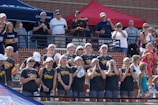 A group of young women, wearing matching Australian team gear, stand enthusiastically in a sports arena. They are smiling, cheering, and clapping, indicating a supportive and lively atmosphere. Spectators in casual attire are visible in the background, some taking photos.