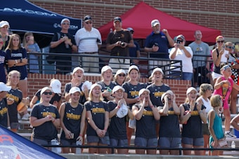 A group of young women, wearing matching Australian team gear, stand enthusiastically in a sports arena. They are smiling, cheering, and clapping, indicating a supportive and lively atmosphere. Spectators in casual attire are visible in the background, some taking photos.