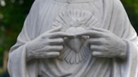 A marble statue depicting a pair of hands gently pointing to a heart symbol on the chest. The heart is encircled by rays, adding a sense of divinity or sacredness. The folds of the robe are meticulously carved, emphasizing grace and tranquility.