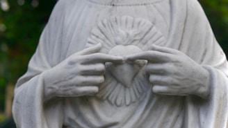 A marble statue depicting a pair of hands gently pointing to a heart symbol on the chest. The heart is encircled by rays, adding a sense of divinity or sacredness. The folds of the robe are meticulously carved, emphasizing grace and tranquility.