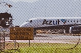 A commercial airplane is taxiing on a runway with 'Azul' branding visible on the side. The aircraft appears to be preparing for takeoff or has just landed, with mountains forming a backdrop in the distance. A wire fence in the foreground and a yellow sign with text in Portuguese indicate an airport environment. The sunny setting and clear visibility suggest a fair weather day.