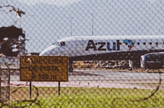 A commercial airplane is taxiing on a runway with 'Azul' branding visible on the side. The aircraft appears to be preparing for takeoff or has just landed, with mountains forming a backdrop in the distance. A wire fence in the foreground and a yellow sign with text in Portuguese indicate an airport environment. The sunny setting and clear visibility suggest a fair weather day.