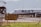 A commercial airplane is taxiing on a runway with 'Azul' branding visible on the side. The aircraft appears to be preparing for takeoff or has just landed, with mountains forming a backdrop in the distance. A wire fence in the foreground and a yellow sign with text in Portuguese indicate an airport environment. The sunny setting and clear visibility suggest a fair weather day.