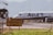A commercial airplane is taxiing on a runway with 'Azul' branding visible on the side. The aircraft appears to be preparing for takeoff or has just landed, with mountains forming a backdrop in the distance. A wire fence in the foreground and a yellow sign with text in Portuguese indicate an airport environment. The sunny setting and clear visibility suggest a fair weather day.