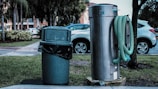 A public outdoor setting featuring a green trash can with a black garbage bag liner next to a large silver utility structure. A green hose is wound around the cylindrical metal object. In the background, several parked cars and trees are visible, with a building partially obscured by foliage.