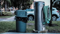 A public outdoor setting featuring a green trash can with a black garbage bag liner next to a large silver utility structure. A green hose is wound around the cylindrical metal object. In the background, several parked cars and trees are visible, with a building partially obscured by foliage.