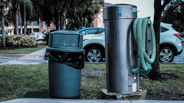 A public outdoor setting featuring a green trash can with a black garbage bag liner next to a large silver utility structure. A green hose is wound around the cylindrical metal object. In the background, several parked cars and trees are visible, with a building partially obscured by foliage.