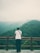 A person wearing a white shirt and dark pants stands on a stone platform, leaning against a wooden railing overlooking a lush, green forest. Mist or clouds partially obscure the distant mountains, creating a serene and contemplative atmosphere.