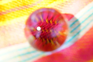 A close-up view of a vibrant, multicolored textile with a glass marble placed on top, creating a blurred effect that distorts the fabric patterns beneath it. The fabric features a blend of yellow, red, and blue stripes, with the marble centering focus on the intricate textures.