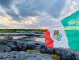 A Mexican flag waves prominently against a scenic coastal landscape. The landscape includes rocky terrain with patches of greenery, leading to a shoreline under a dramatic cloudy sky.