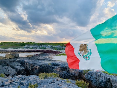A Mexican flag waves prominently against a scenic coastal landscape. The landscape includes rocky terrain with patches of greenery, leading to a shoreline under a dramatic cloudy sky.