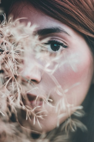A close-up of a person's face partially obscured by delicate branches or dried plants. The focus is on the eye, which is highlighted with makeup, while the rest of the face has soft, natural tones. The image seems to convey a sense of mystery and introspection.