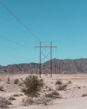 A dry, sandy desert landscape with sparse vegetation and several tall electrical power lines running through it. In the background, there is a range of rocky mountains under a clear blue sky.