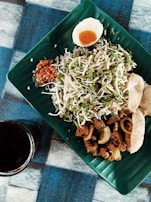 A beautifully arranged meal on a green, leaf-shaped plate featuring sliced vegetables, a hard-boiled egg cut in half, spicy sauce, fried meat pieces, and crispy crackers. A dark beverage in a glass is placed next to the plate on the blue and black checkered tablecloth.