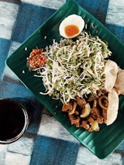A beautifully arranged meal on a green, leaf-shaped plate featuring sliced vegetables, a hard-boiled egg cut in half, spicy sauce, fried meat pieces, and crispy crackers. A dark beverage in a glass is placed next to the plate on the blue and black checkered tablecloth.