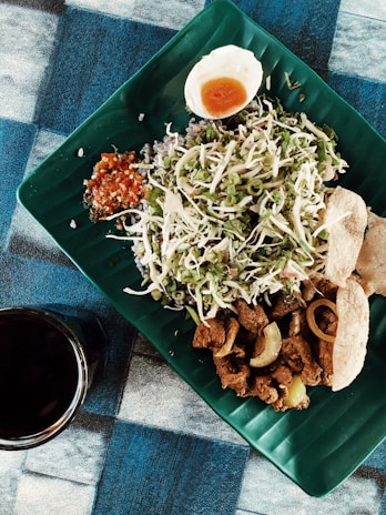 A beautifully arranged meal on a green, leaf-shaped plate featuring sliced vegetables, a hard-boiled egg cut in half, spicy sauce, fried meat pieces, and crispy crackers. A dark beverage in a glass is placed next to the plate on the blue and black checkered tablecloth.